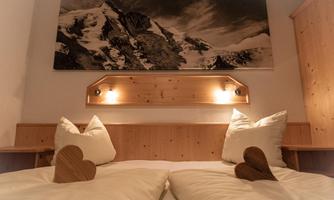 A cozy bedroom with wooden decor and two heart-shaped pillows. On the wall hangs a landscape painting with mountains and snow.