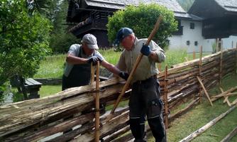 Two men are building a wooden fence in a rural setting. In the background, old buildings and plenty of green nature can be seen.