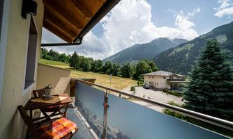 A balcony with two chairs and a table, surrounded by mountains and trees. The sky is clear with a few clouds.