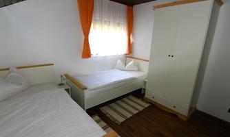A simple bedroom with two single beds and a white wardrobe. Thick curtains and a warm wooden floor create a cozy atmosphere.