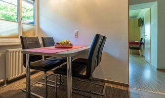 A bright dining area with a table and four black chairs. A fruit bowl is on the table, and a room is visible in the background.
