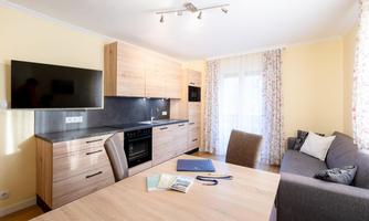 A modern kitchen with wooden cabinets and a television on the wall. The room features a table with chairs and a cozy sofa.