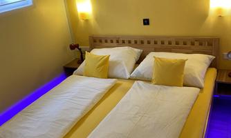 A cozy bedroom with a large double bed and yellow pillows. The walls are painted yellow, and there is atmospheric lighting under the bed.