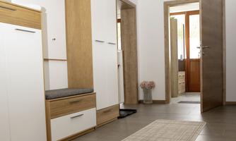 A modern hallway with wooden and white furniture. The door leads to another room, where a bright, inviting space can be seen.