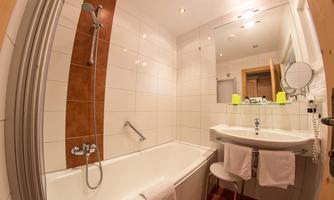 A modern bathroom with a bathtub and a shower. The room is bright and minimally furnished, with a large mirror and a sink.
