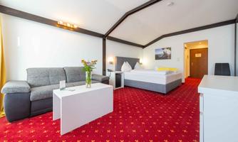 A cozy hotel room with red carpeting. There is a sleeping area with a large bed and a seating area with a gray sofa and a table.