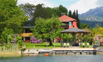 A picturesque house by the lake with green trees and colorful flowers. A terrace with a sun umbrella invites you to relax.