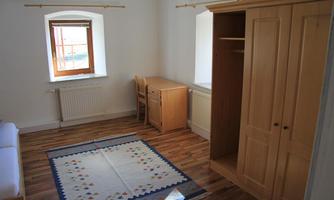 A simple room with a light wood floor. It has a window, a table, a chair, and a wardrobe.