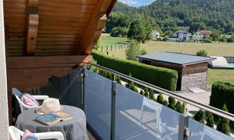 A cozy balcony with a table and chairs, surrounded by a beautiful landscape. In the background, mountains and a blue sky can be seen.
