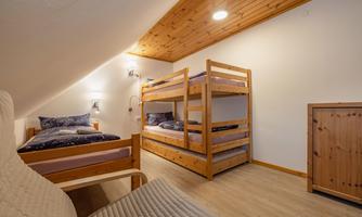 A cozy bedroom with wooden bunk beds and a single bed. The walls are light, and the ceiling has an appealing wood paneling.