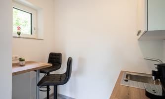 A modern kitchen corner with a small wooden bar and two black chairs. The window lets in plenty of light and a plant decoration creates an inviting atmosphere.