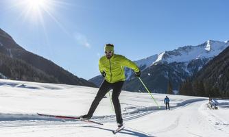 An active cross-country skier in a bright yellow jacket is skiing through a snowy landscape. In the background, there are mountains and a bright blue sky.