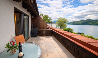 A cozy terrace with a table and chairs offers a view of the lake and the surrounding landscape. Sky with clouds complements the pleasant atmosphere.