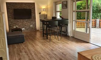 A modern living room with wooden flooring and a cozy seating area. There is a dining table and large windows that let in plenty of daylight.