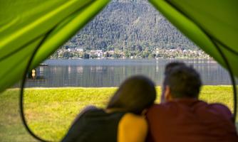 A couple relaxes in the tent and gazes at a peaceful lake. In the background, green hills and a small town can be seen.