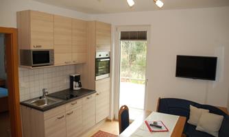 A modern kitchen with wooden cabinets and white tiles. The room is bright and friendly furnished, with a table and a sofa.