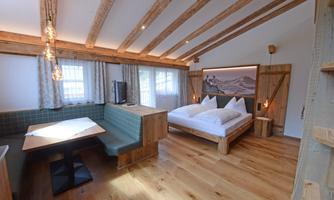 A modern, cozy room with wooden ceilings and a large bed. There is a dining area with a corner bench and plenty of daylight from the windows.