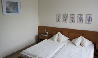 A simple bedroom with a double bed and two pillows. Pictures hang on the wall, and there is a nightstand with a bottle of water.