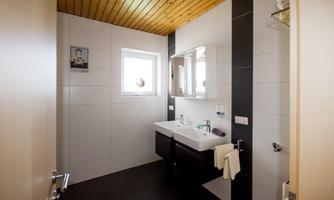A modern bathroom with a light wooden ceiling and white walls. It features a double sink and a window that lets in daylight.