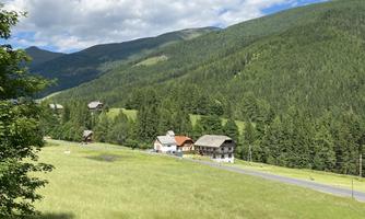A picturesque landscape with gentle hills and wooded mountains. In the foreground, several traditional houses and green meadows can be seen.