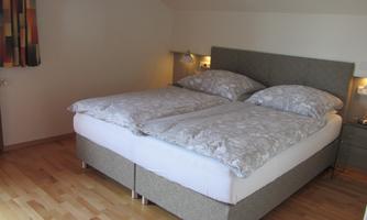 A cozy bedroom with a double bed and light wooden floor. The walls are plain, and there is window decoration in soft colors.