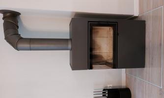A modern wood stove made of black metal stands against a wall. The chimney flue is visible and the floor is covered with wooden tiles.