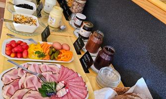 A hearty breakfast buffet with cold cuts, cheese, fresh fruits, and rolls. There is also jam, yogurt, and various snacks.