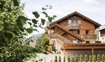 A modern wooden house with a balcony and windows. Surrounded by trees and green plants.