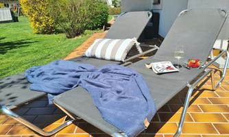 Two loungers on a terrace with a blue towel and a cushion. On a table, there is a book and a glass with a drink.