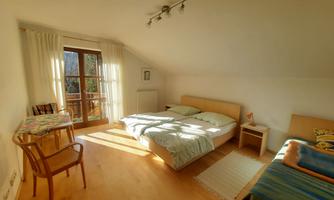 A cozy bedroom with two beds and a table. The room has large windows through which plenty of daylight streams.