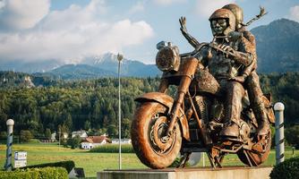 A large statue of a couple on a motorcycle, joyfully riding. In the background, mountains and a green landscape can be seen.