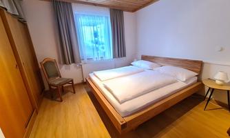 A simple room with two beds and a wooden floor. There is a window with curtains and a small table next to the bed.