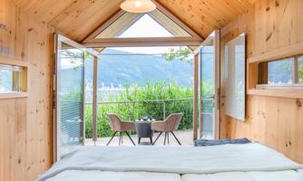 A cozy wooden accommodation with a view of nature. In the foreground, a large bed and a balcony with two chairs and a table can be seen.