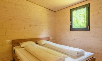 A simple bedroom with wooden walls and a large bed. The view from the window shows the green surroundings.