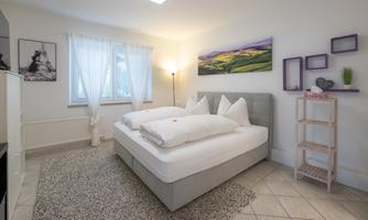 A cozy bedroom with a large bed, two pillows, and soft lighting. There are windows with curtains and some decorative elements on the wall.