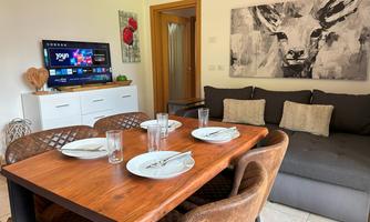 A cozy living area with a dining table, chairs, and a green television. A modern artwork hangs on the wall, and there is a comfortable couch.
