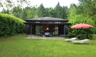 A modern house with large windows in the midst of a green garden. On the terrace, there are chairs and a sun umbrella.