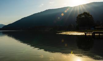 A tranquil lake at the foot of a mountain, surrounded by trees. The sun shines gently and reflects on the water's surface.