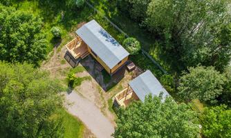 Two cozy wooden cabins surrounded by green trees and meadows. The path leads through nature to the cabins.