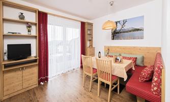A bright living room with a dining area and a cozy seating corner. The walls are in neutral colors, and large windows provide plenty of daylight.