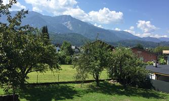 A picturesque landscape with green meadows, trees, and mountains in the background. The sky is clear and blue, creating a sunny atmosphere.