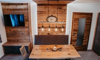 A modernly furnished dining area with a large wooden table and chairs. The walls are made of wood, and there is cozy lighting as well as a television.
