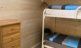 A simple room with a bunk bed and a wooden chest of drawers. The walls are made of wood and the floor is tiled.