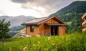 A cozy wooden house surrounded by green meadows and gentle hills. The sun shines through the clouds, creating an inviting atmosphere.