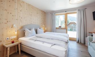 A cozy bedroom with wooden paneling and a large window. The room is bright and modernly furnished, with a large bed and a small bedside table.