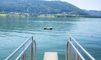 A tranquil lakescape with a pier leading into the water. In the background, gentle hills and a clear blue sky can be seen.