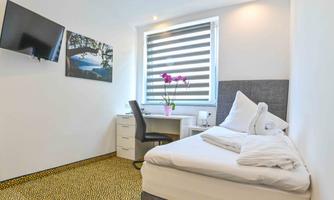 A modern room with a single bed, a desk, and a television. A beautiful orchid stands on the table in front of a window with blinds.