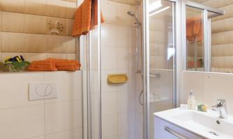 A modern bathroom with a glass shower and a sink. The walls are light and paneled with wood, and there are orange towels.