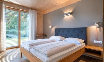A modern bedroom with a cozy double bed and wooden flooring. Large windows let in plenty of daylight and offer a view of nature.