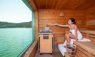 A modern sauna with windows that offer a view of a tranquil lake. A woman is sitting relaxed in the sauna and enjoying the atmosphere.